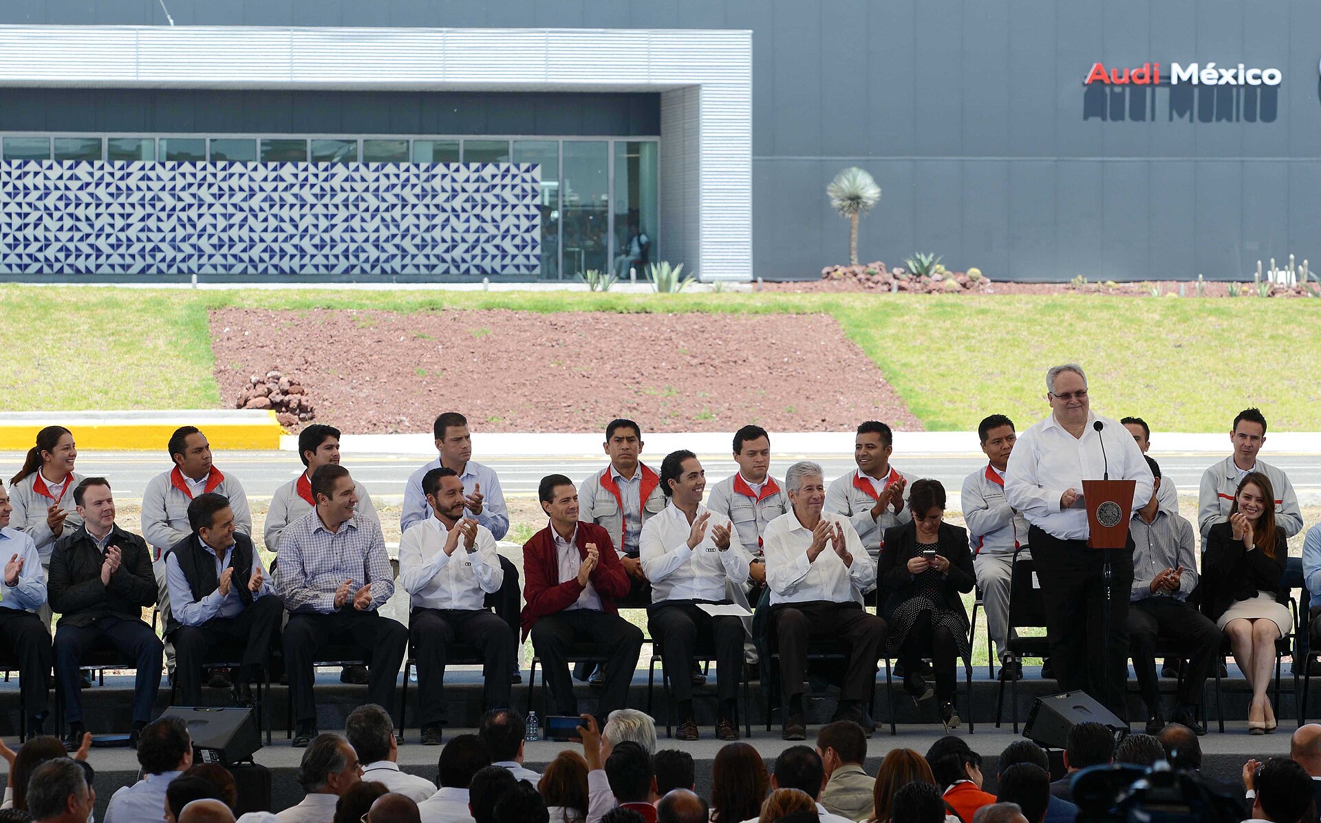 Rows of seated officials and workers in uniforms applauding in front of the Audi México plant building in San José Chiapa, Puebla