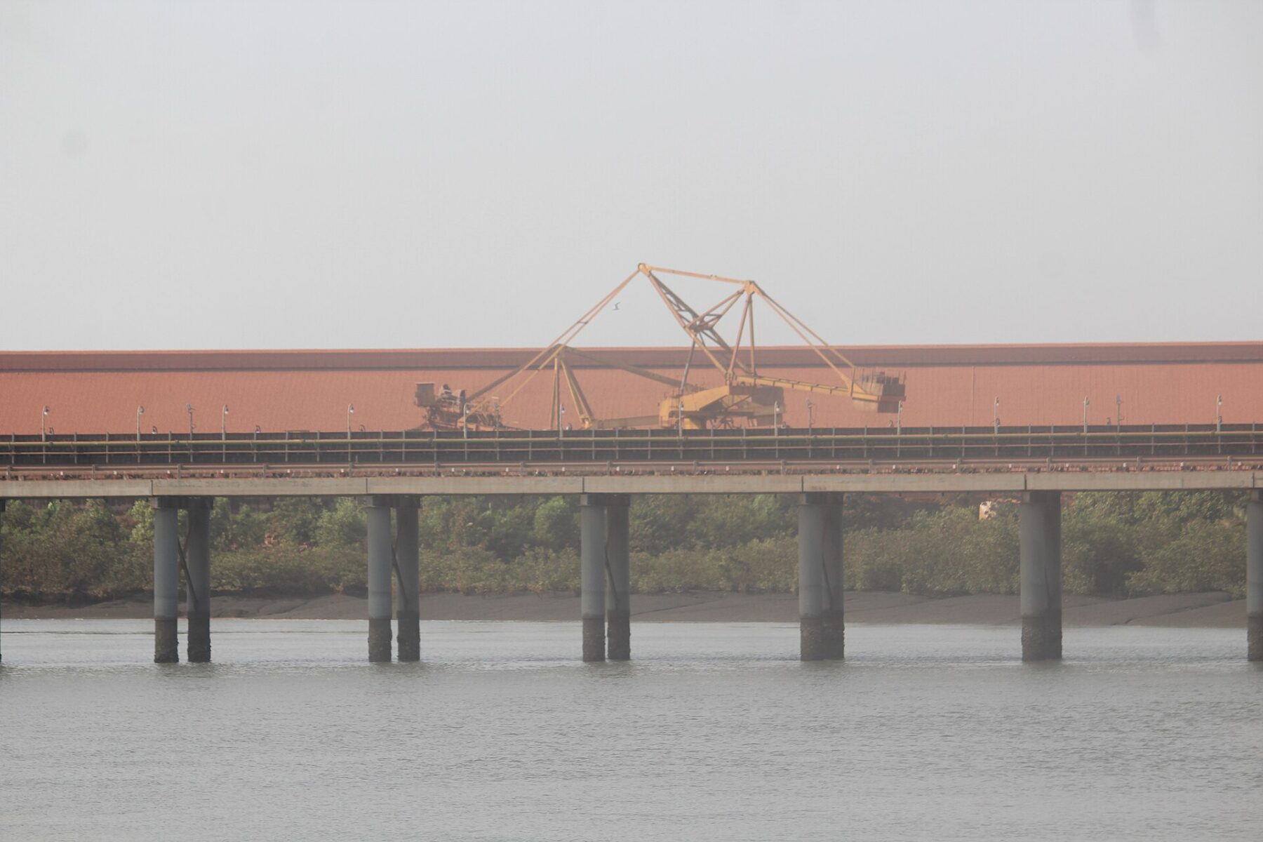 An elevated conveyor on concrete pillars crosses an estuary in front of a long red-roofed bauxite storage shed with a yellow stacker machine