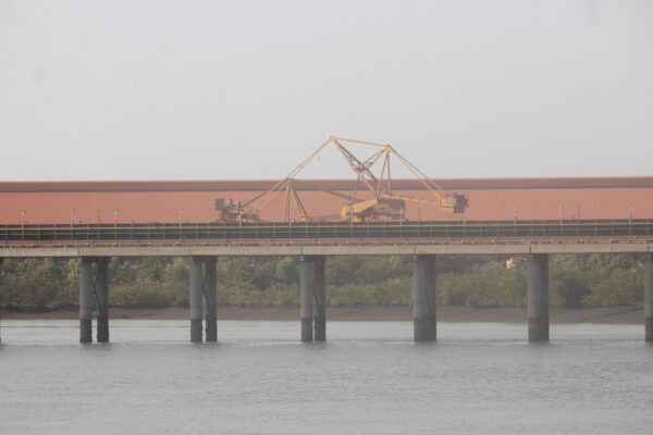 An elevated conveyor on concrete pillars crosses an estuary in front of a long red-roofed bauxite storage shed with a yellow stacker machine