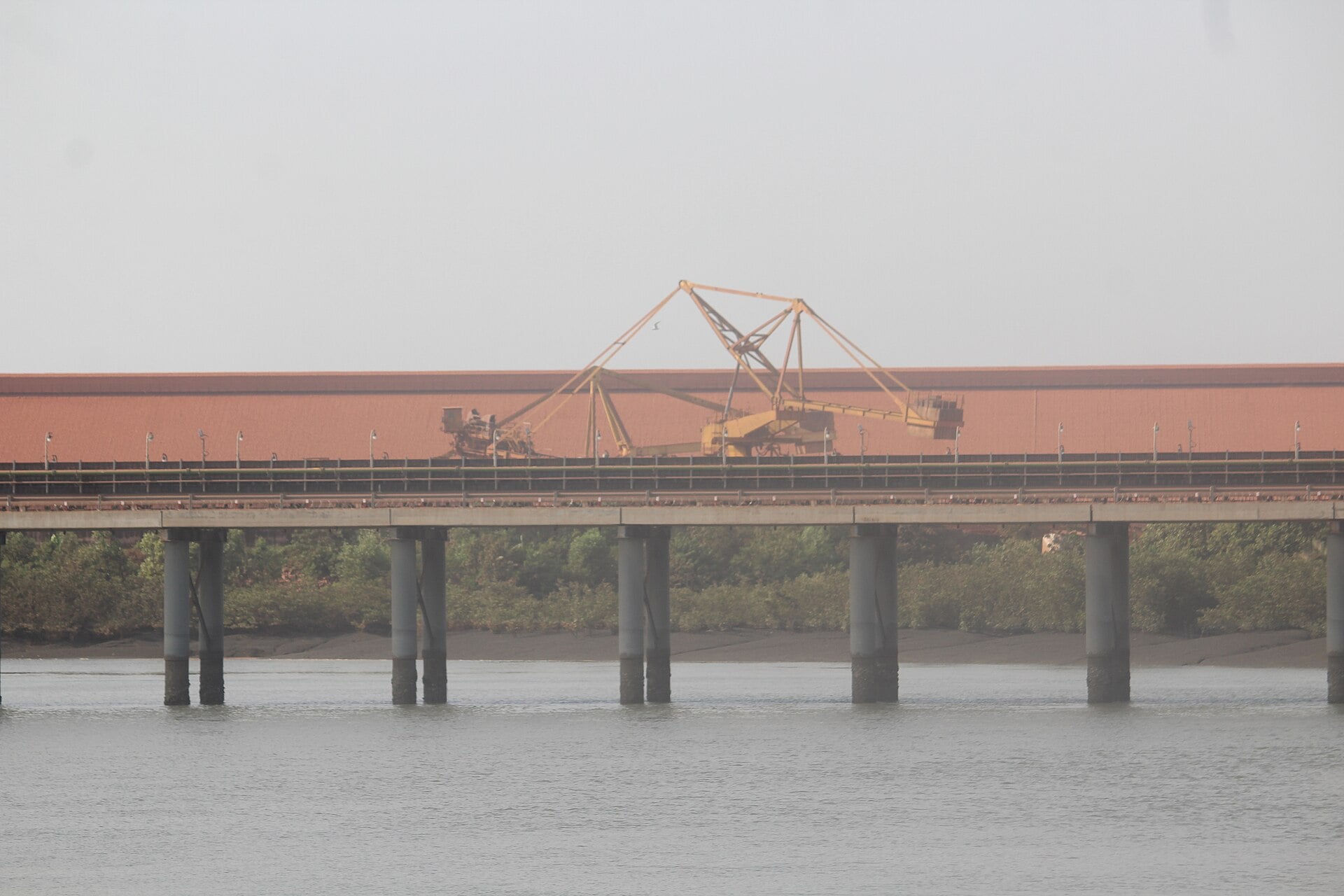An elevated conveyor on concrete pillars crosses an estuary in front of a long red-roofed bauxite storage shed with a yellow stacker machine