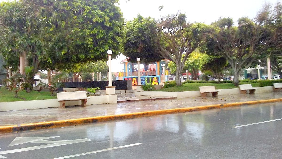 Town park with benches, trees and a colourful sign spelling Nagua beside a wet road in Nagua, Dominican Republic