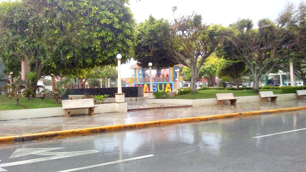 Central plaza in Nagua, Dominican Republic, with colourful letters spelling the town's name, trees, benches and a wet street