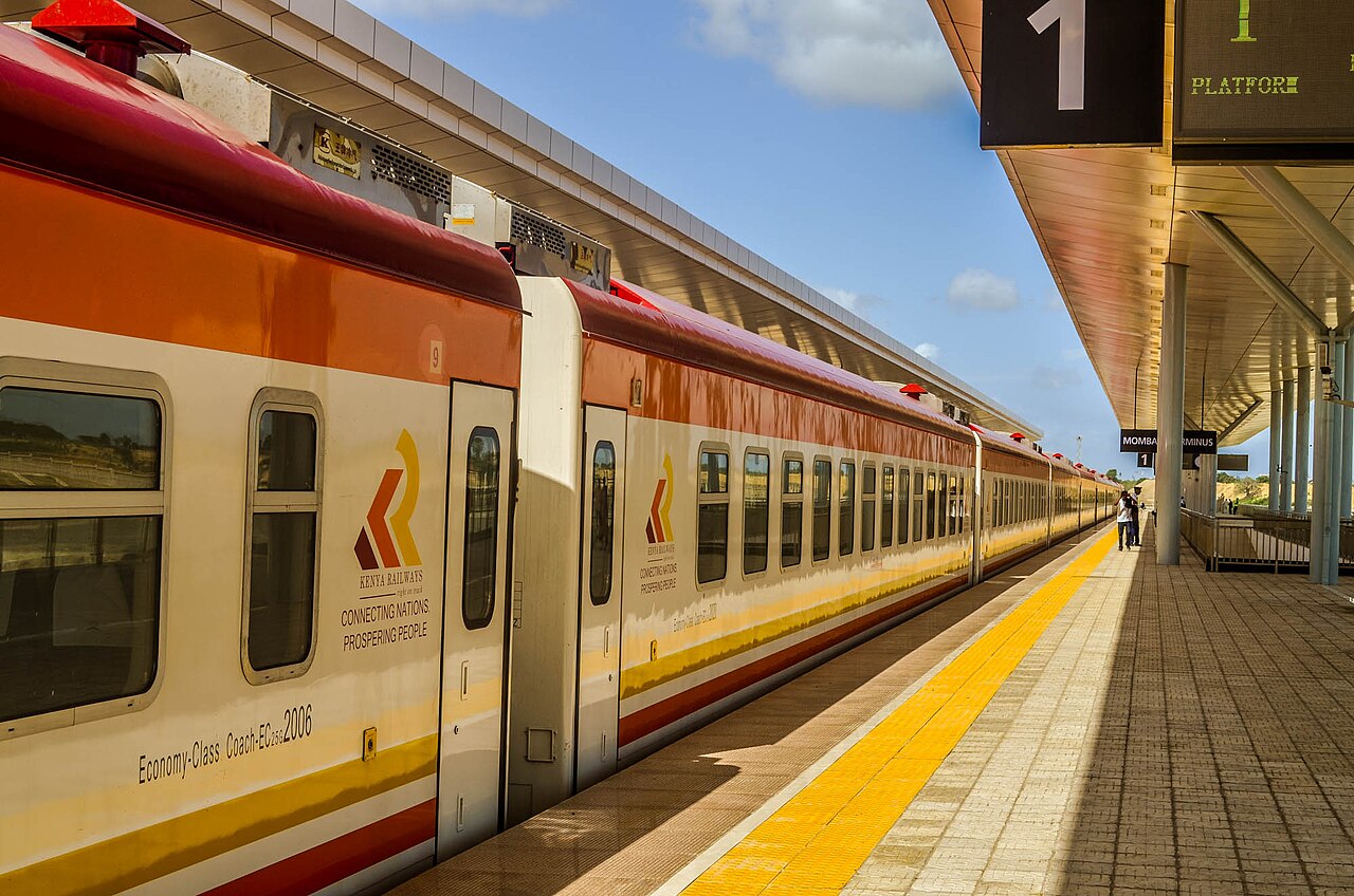 An orange and cream Kenya Railways Madaraka Express economy coach standing at a station platform under a long canopy.
