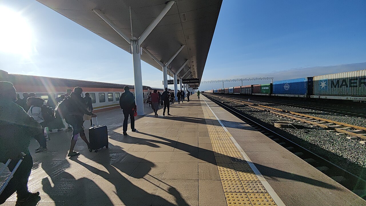Passengers with luggage walk along a covered platform at Nairobi Terminus railway station with a train on the left and freight containers on the right.