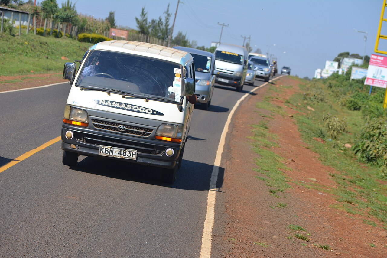 A white Toyota minibus used as a matatu on a paved road in Kenya, with other vehicles queuing behind it