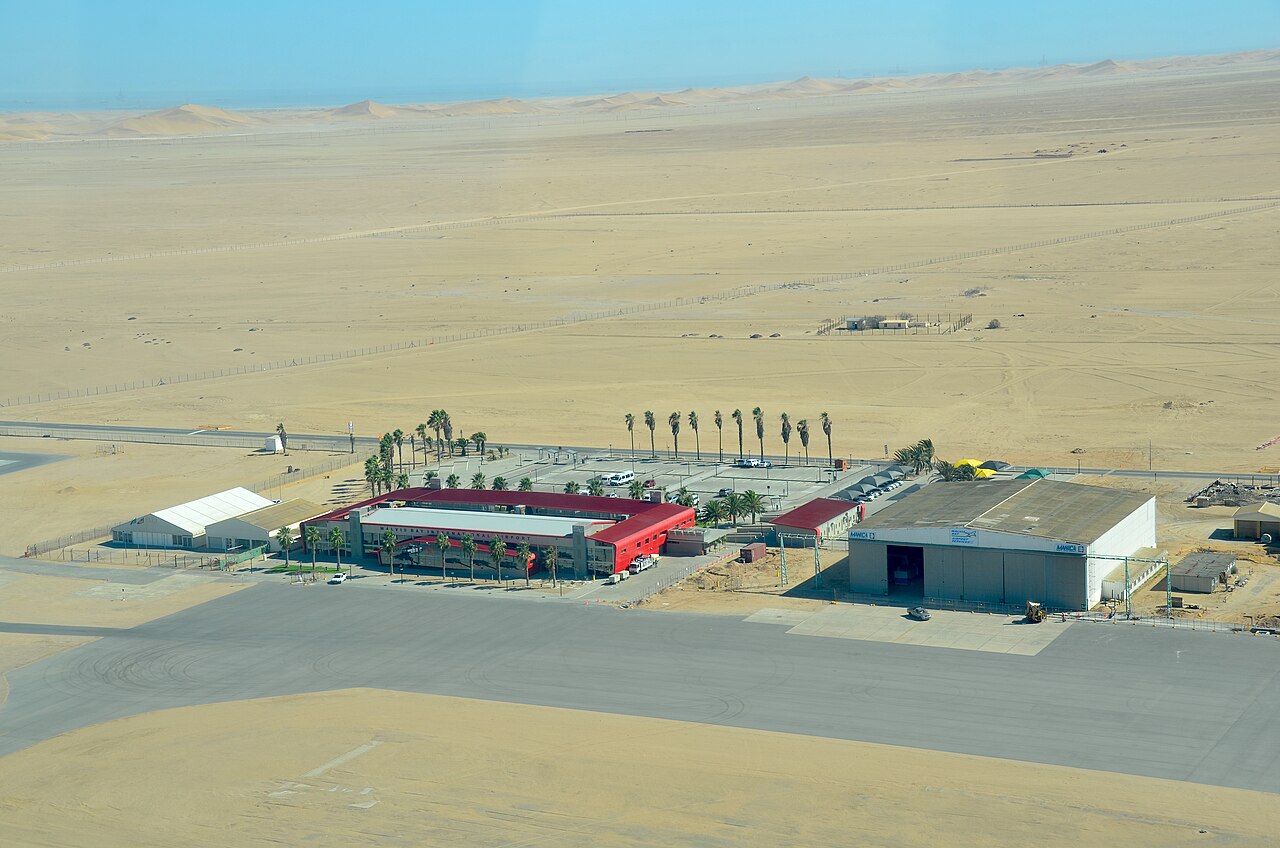 Aerial view of the red-roofed terminal and a hangar of Walvis Bay Airport on flat desert, with sand dunes behind.