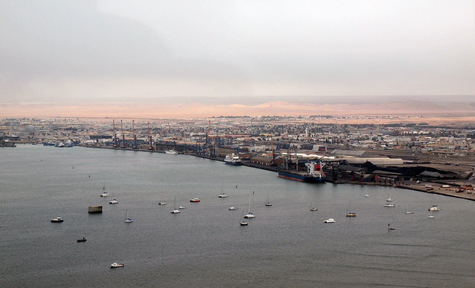 The port of Walvis Bay seen from the air, with ships at the quay, container cranes, sailing boats on the bay and desert dunes behind the town