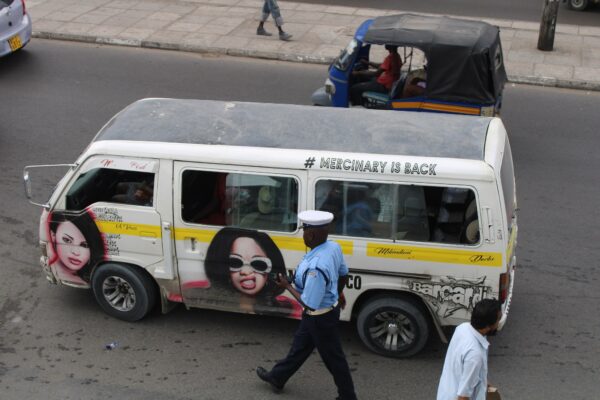 A Kenya Police officer in a pale blue shirt and white cap walks past a decorated minibus on a busy road