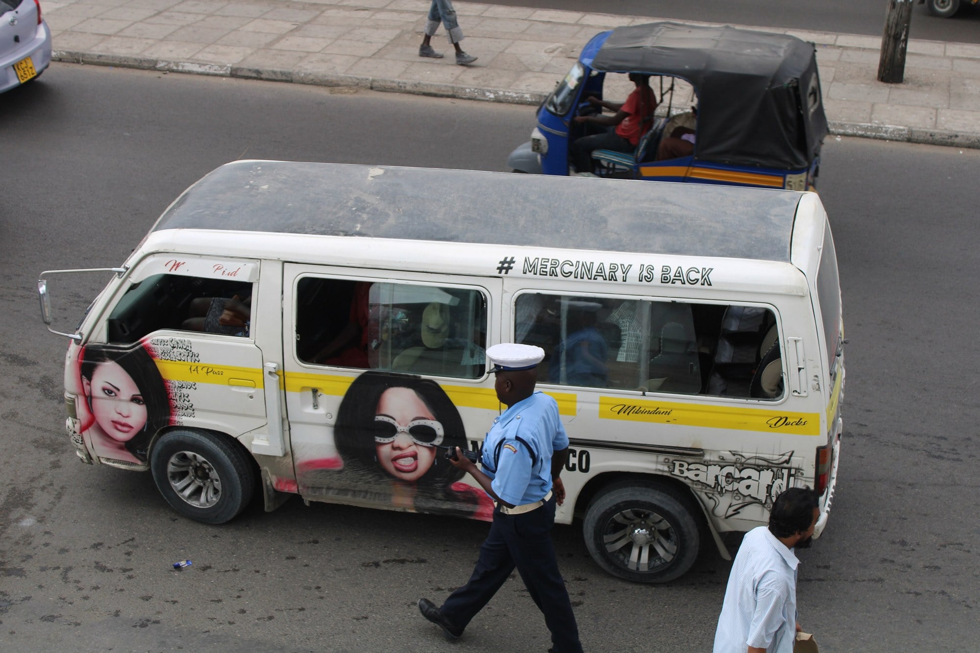 A Kenya Police officer in a pale blue shirt and white cap walks past a decorated minibus on a busy road