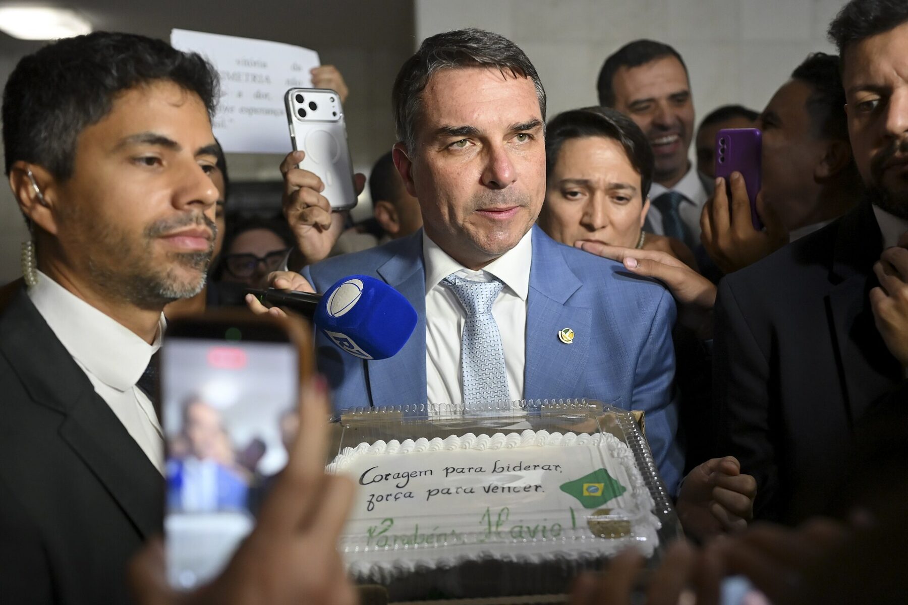 Senator Flávio Bolsonaro in a blue suit and light tie among reporters and aides, with a microphone at his chest and a cake in front of him