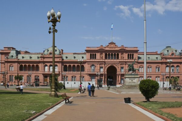 The pink Casa Rosada government palace in Buenos Aires on a sunny day, with an Argentine flag above it and people crossing the plaza in front