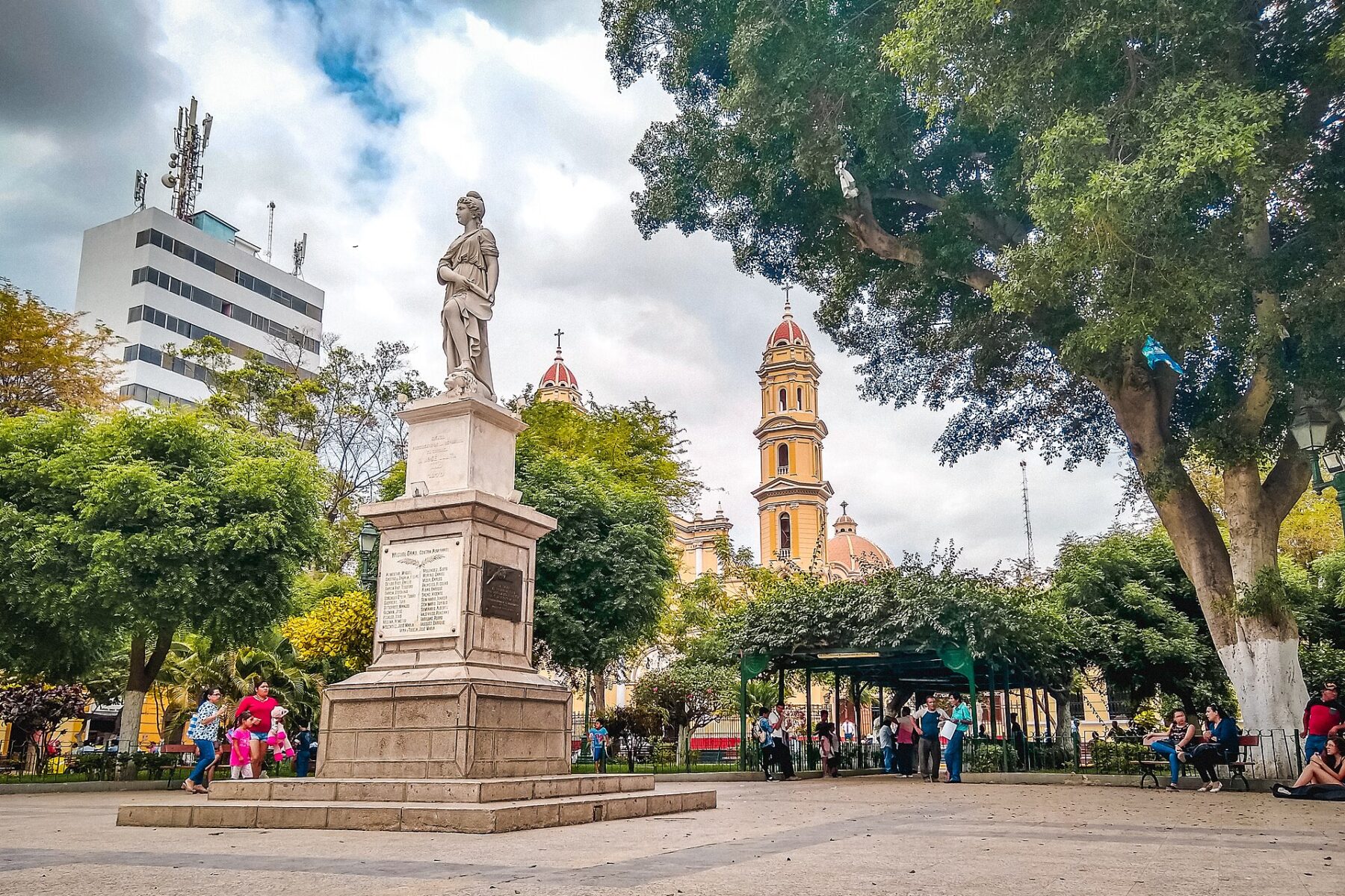 The Plaza de Armas in Piura, Peru, with a white statue on a stone pedestal, green trees and a yellow church tower under a cloudy sky