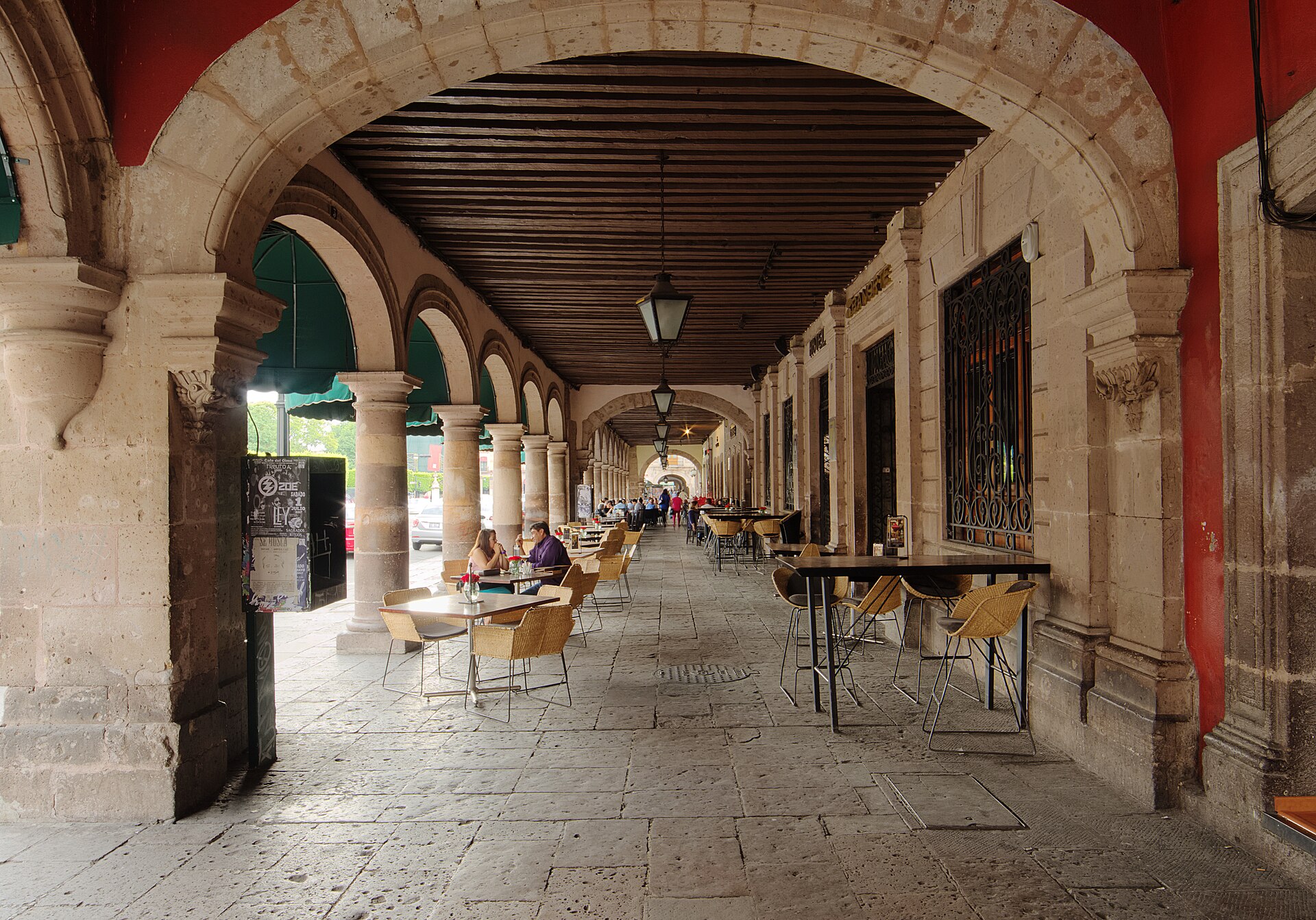 A stone arcade with wooden beams and lanterns in the historic centre of Morelia, with people seated at cafe tables