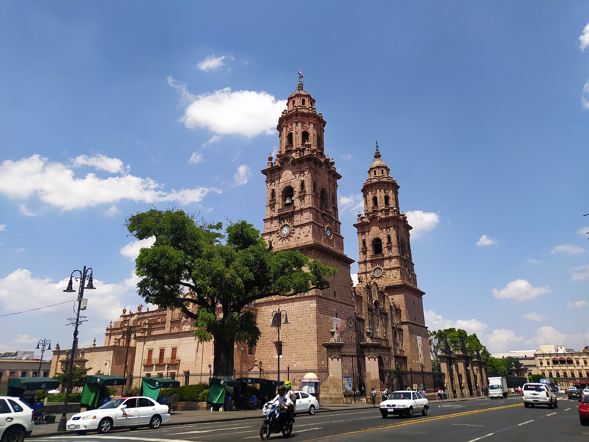 The twin pink-stone towers of Morelia Cathedral behind a green tree on a sunny day, with cars and a motorcycle on the street in front