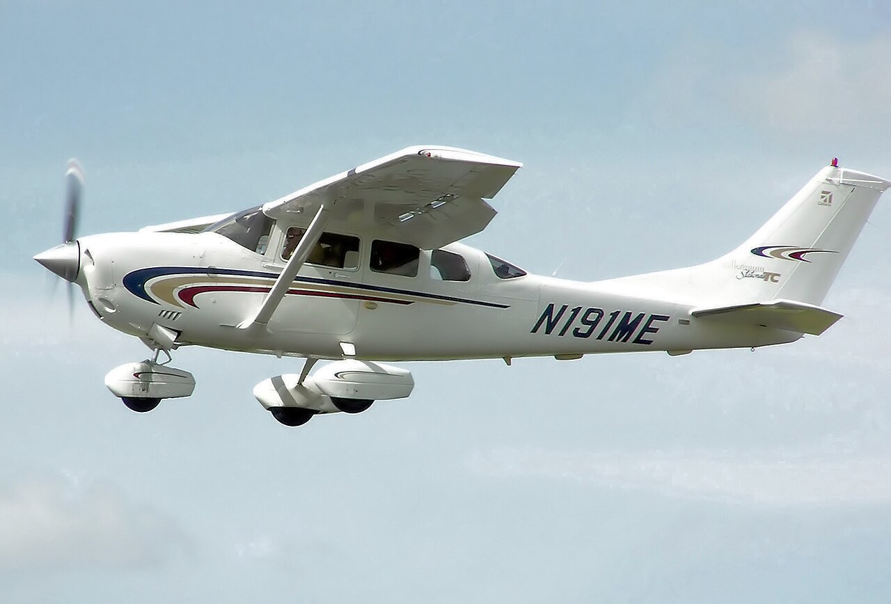 A white Cessna 206H Stationair single-engine light aircraft with blue and red stripes in flight against a pale sky