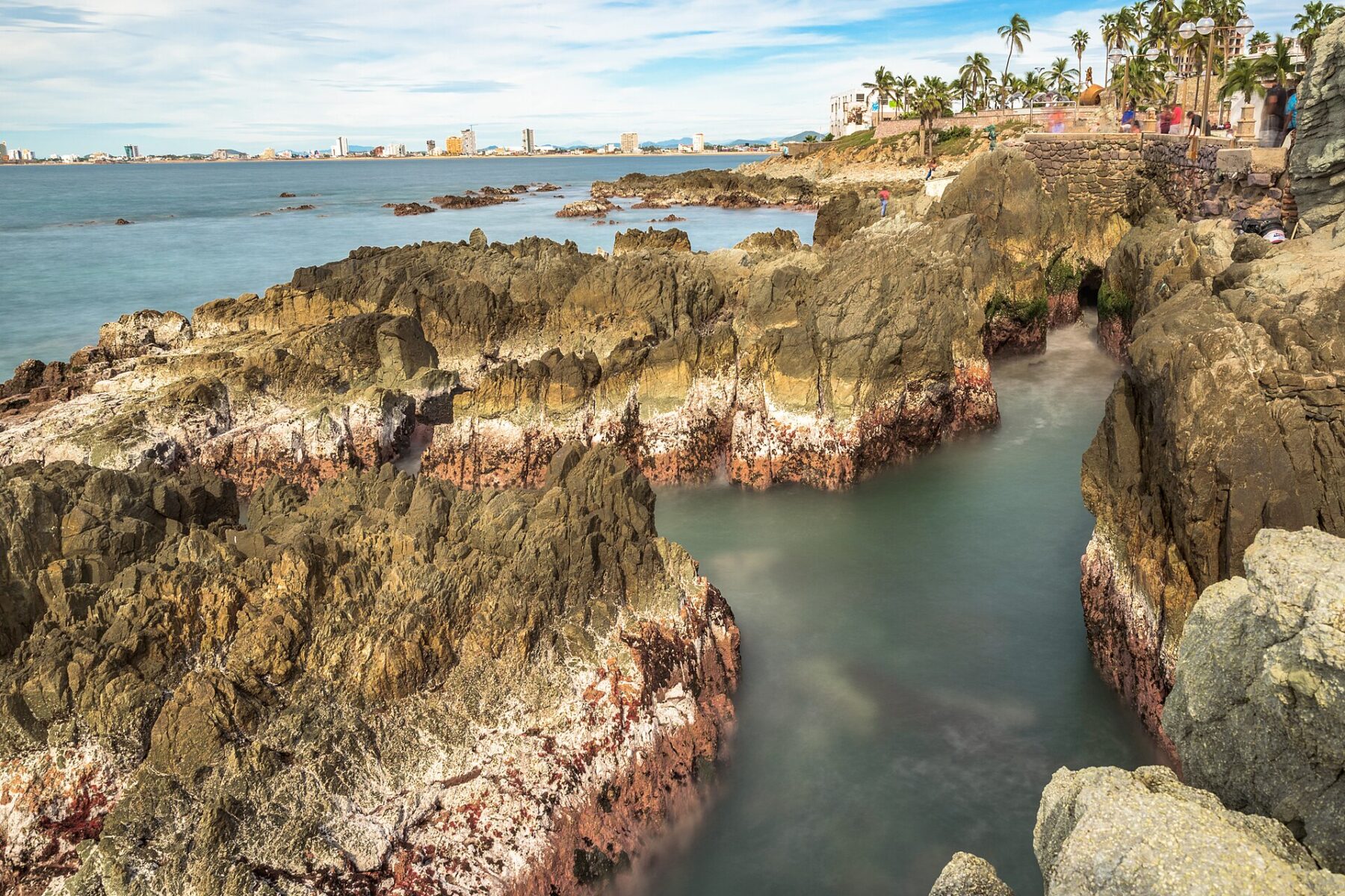 The rocky Pacific coast of Mazatlán, Mexico, with beach-front buildings along the shore in the distance