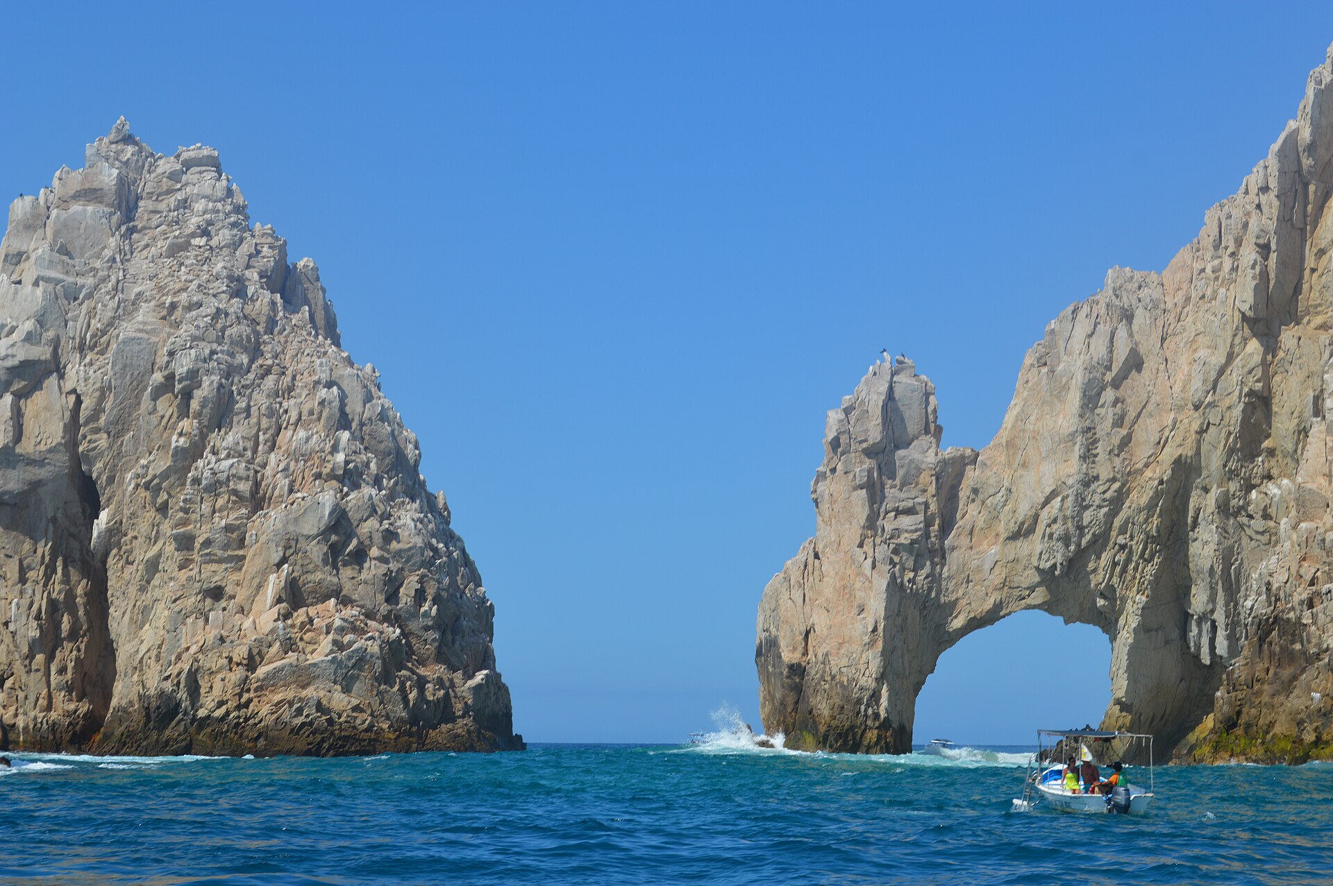 A rock arch rising from the sea at Cabo San Lucas, with a small boat passing in front under a clear blue sky