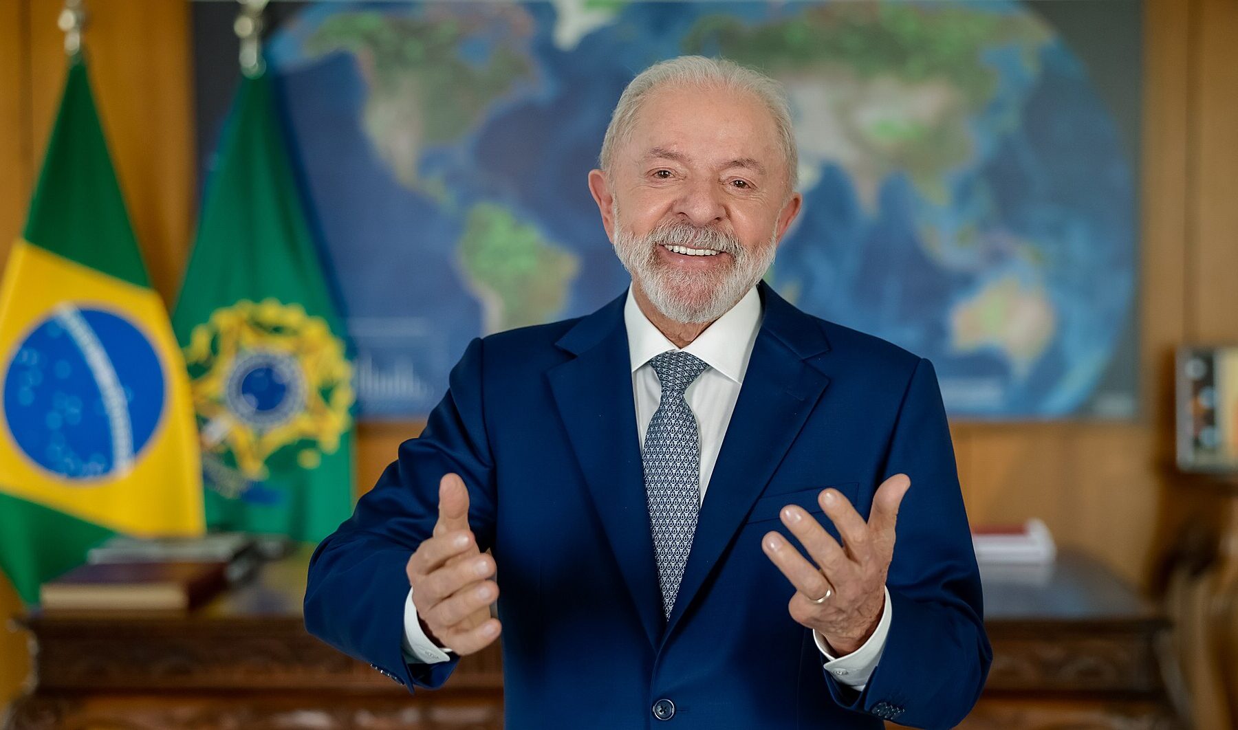 Brazilian President Luiz Inácio Lula da Silva, in a blue suit, gestures with both hands in front of a Brazilian flag and a world map