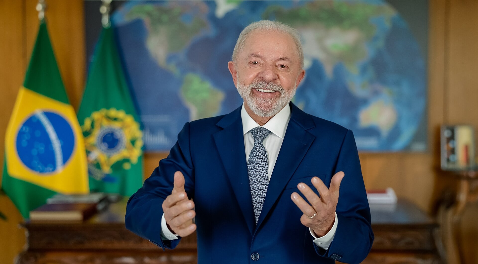 Brazilian President Luiz Inácio Lula da Silva, in a blue suit, gestures with both hands in front of a Brazilian flag and a world map