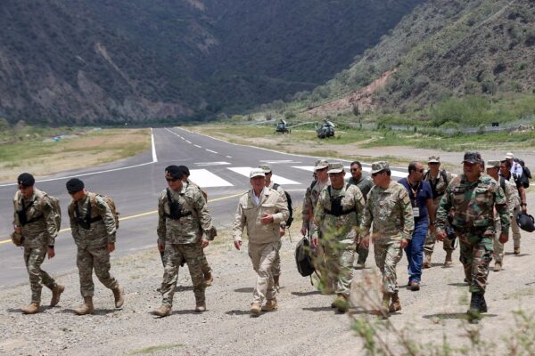 Soldiers in camouflage and officials in civilian clothes walk along a road beside a helicopter pad in a mountain valley