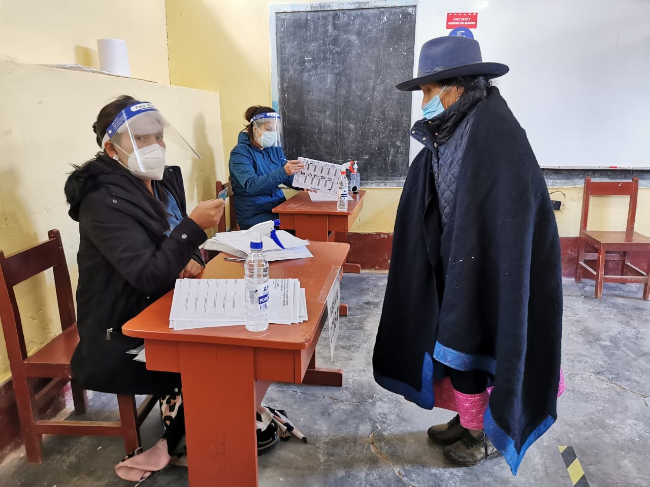 A voter in a dark poncho and blue hat stands at a table where two masked electoral workers in face shields hold ballot papers