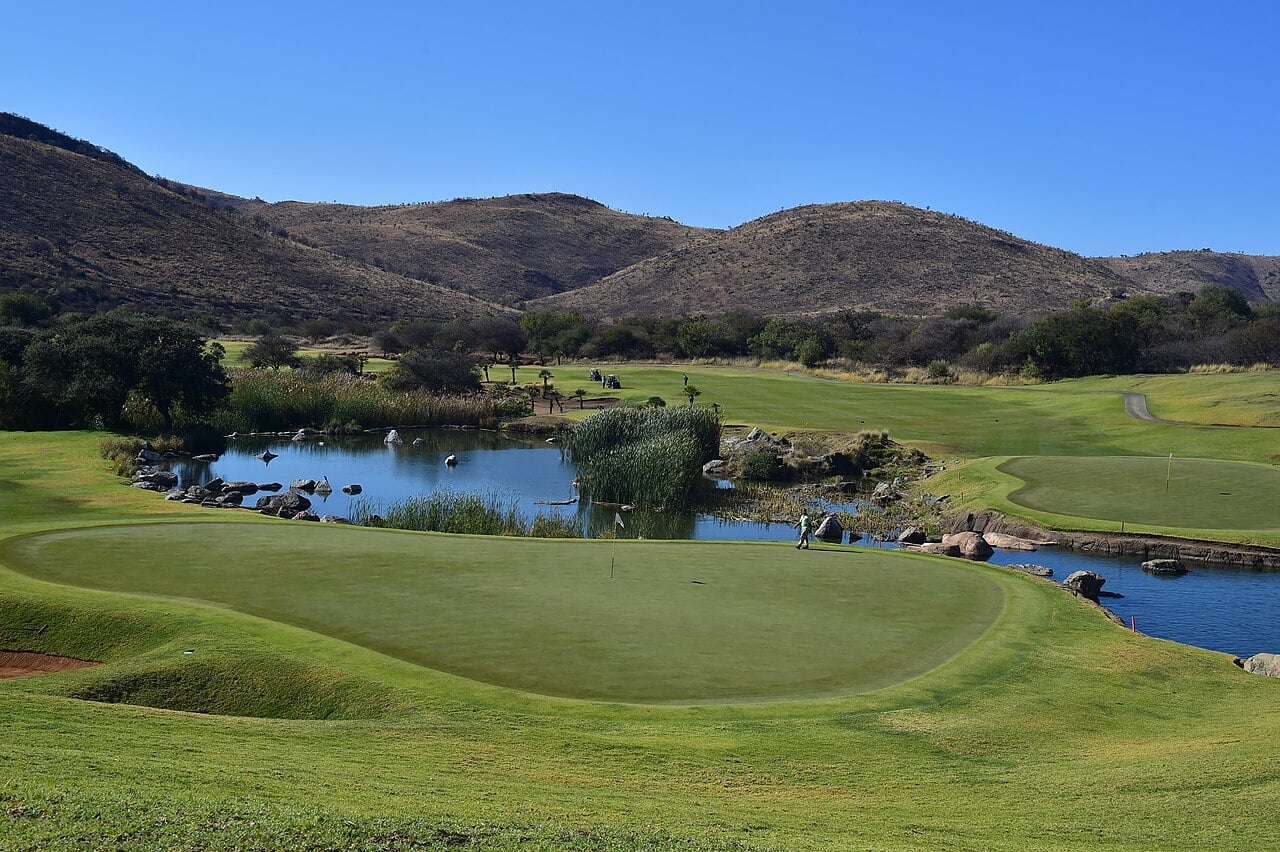 A golf green beside a reed-lined pond with boulders, a fairway beyond and dry brown hills under a clear blue sky at Sun City in South Africa