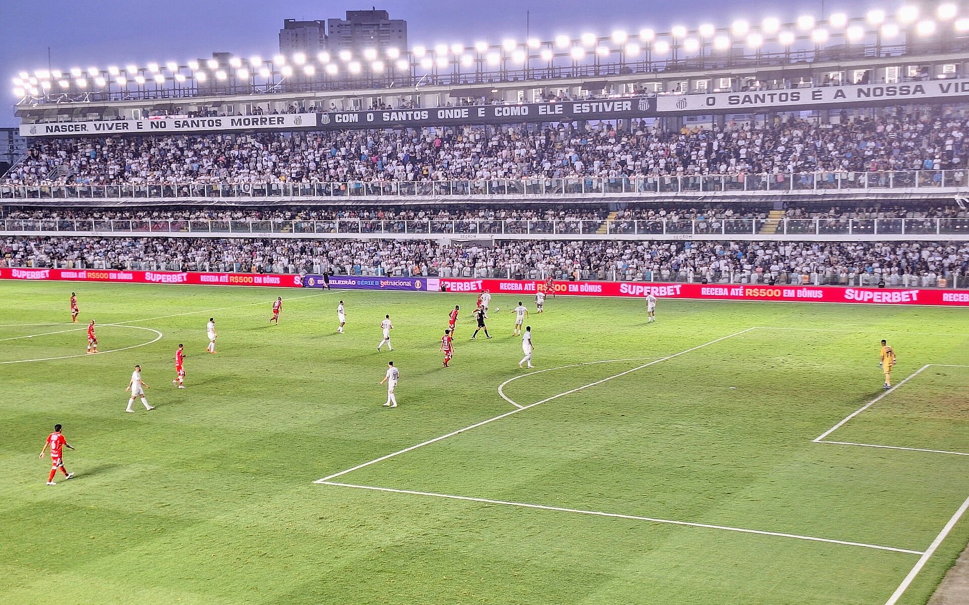 Players in white and red on the Vila Belmiro pitch under floodlights at dusk, with packed stands and advertising boards behind them