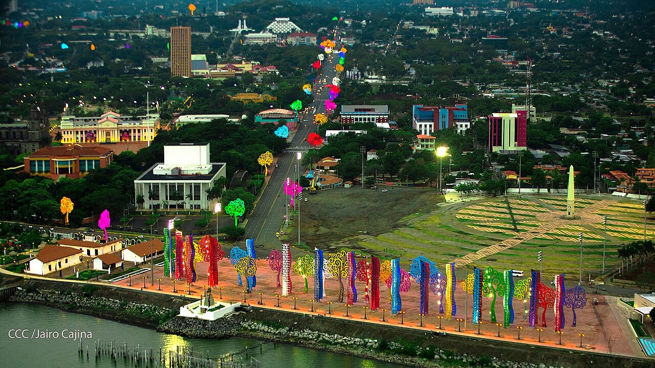 Aerial view of central Managua at dusk, with colourful tree-shaped sculptures along the waterfront and government buildings behind.