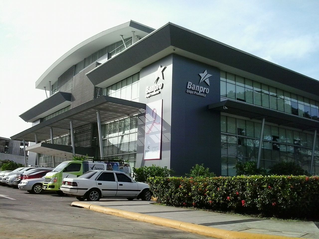 Modern office building with a curved roof and the Banpro Grupo Promerica name on its walls, with cars parked in front, in Managua.