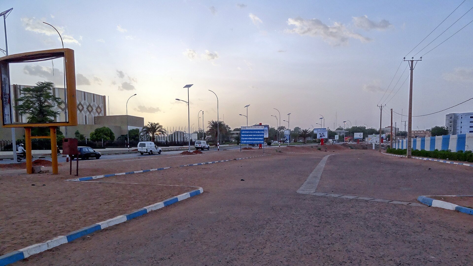 A wide boulevard in Niamey, the capital of Niger, at dusk with street lamps and passing vehicles