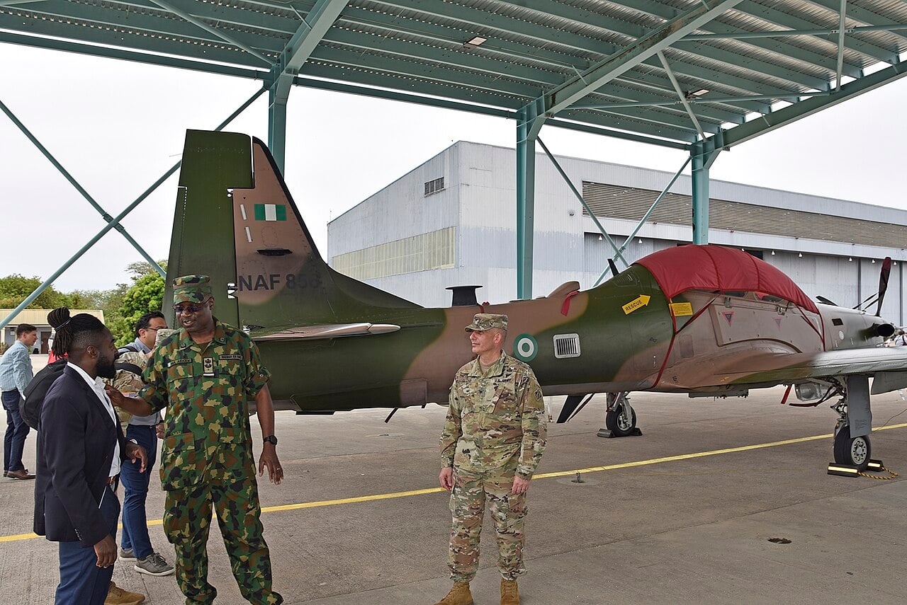 Nigerian and US officers talk beside a camouflaged Nigerian Air Force A-29 Super Tucano plane under a sunshade at Kainji Air Force Base