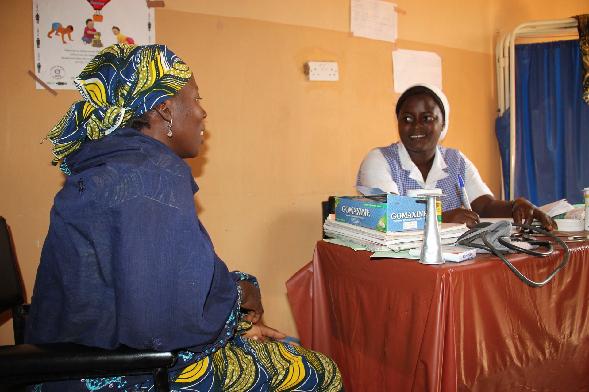 A health worker in a white cap talks with a woman patient across a desk at a clinic in northern Nigeria