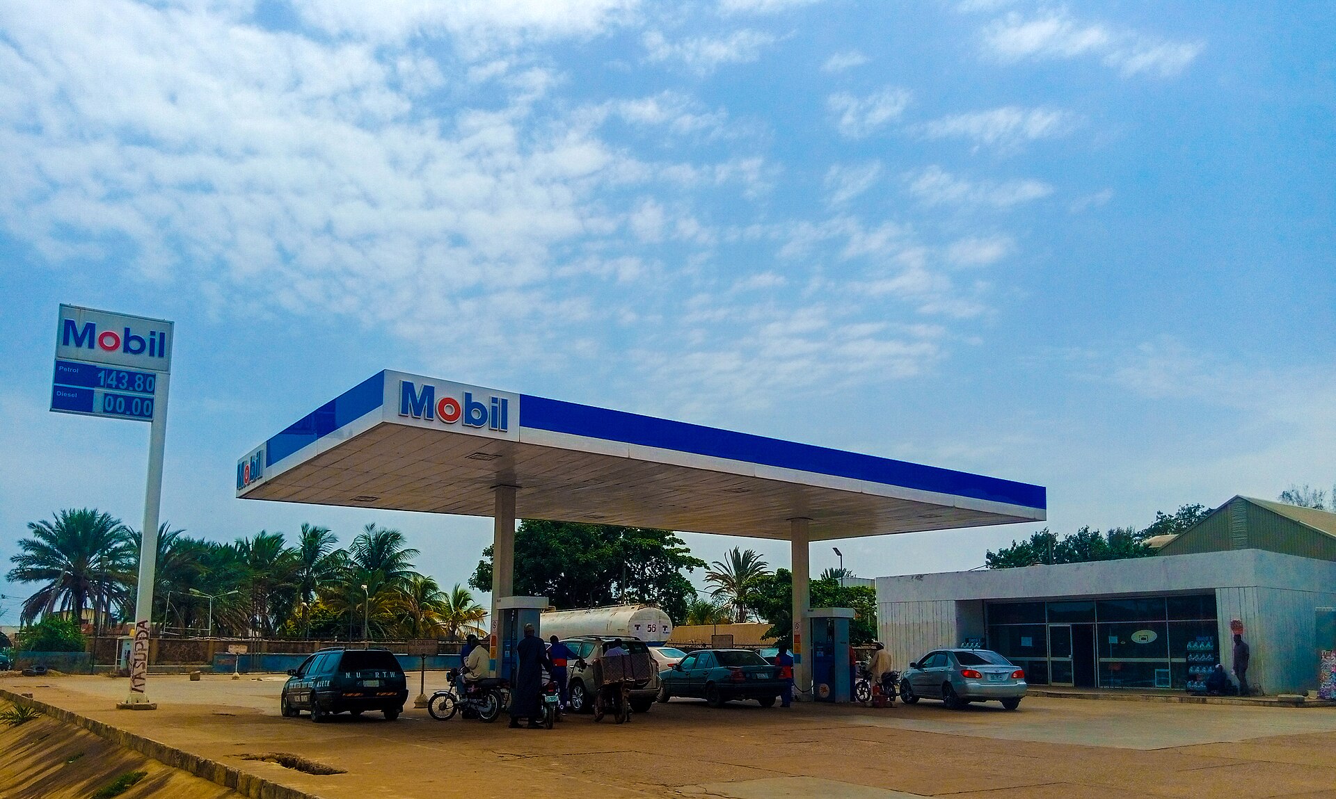 A Mobil-branded filling station with a blue and white canopy, cars and motorbikes on its forecourt, in Nigeria