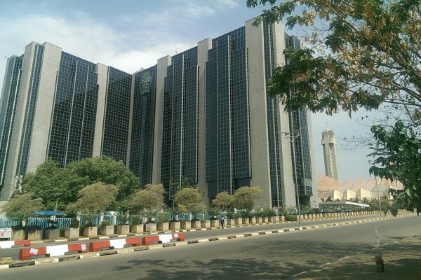 Street-level view of the Central Bank of Nigeria building in Abuja behind trees, with red and white road barriers in front