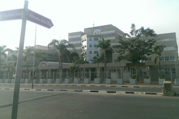 The Federal Ministry of Finance headquarters in Abuja, a beige concrete office block behind palm trees and a fence, with street signs for Ahmadu Bello Way