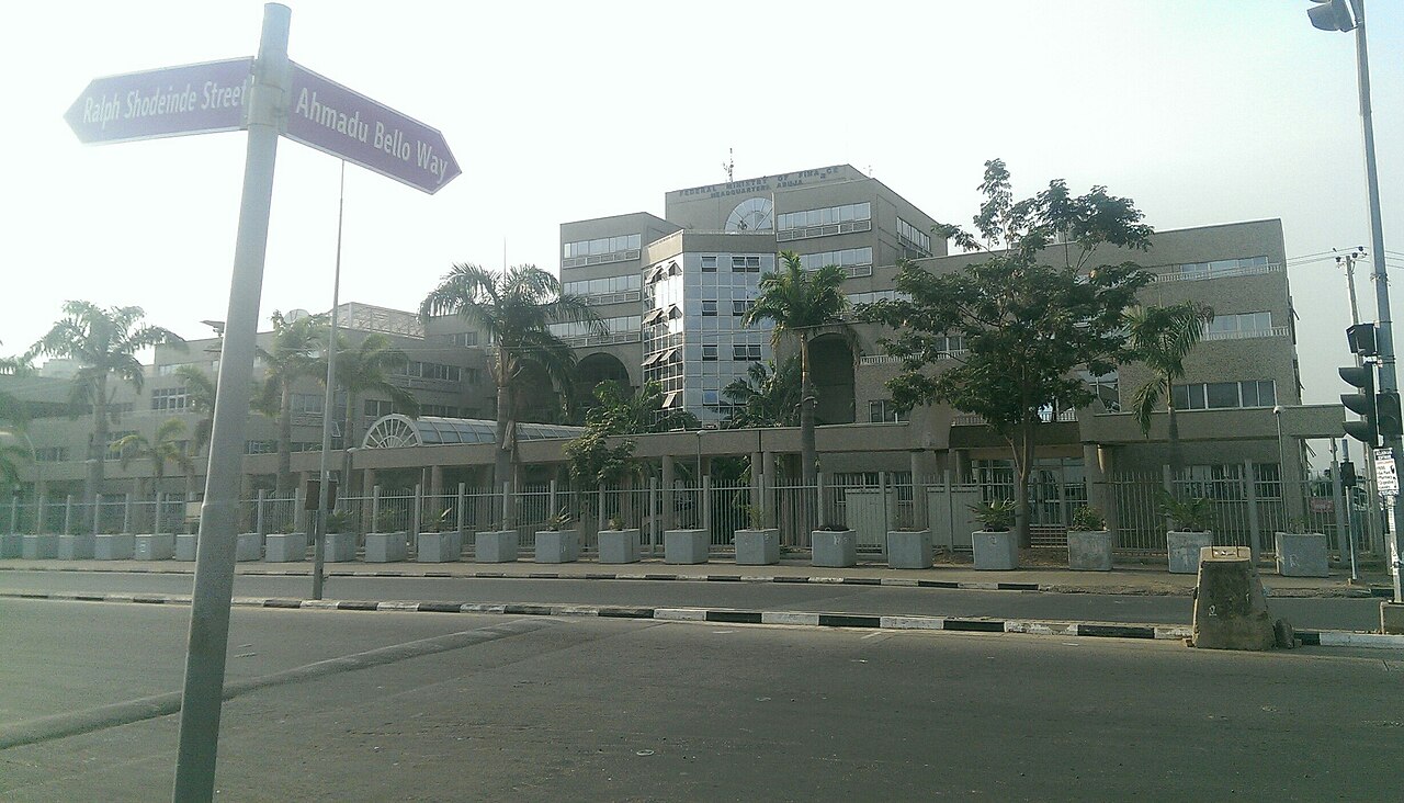 The Federal Ministry of Finance headquarters in Abuja, a beige concrete office block behind palm trees and a fence, with street signs for Ahmadu Bello Way