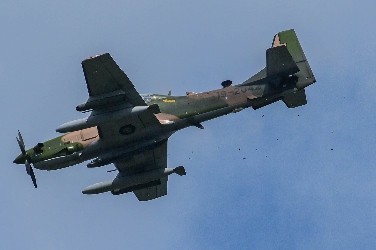 A camouflaged Nigerian Air Force A-29 Super Tucano light attack aircraft firing its guns against a blue sky
