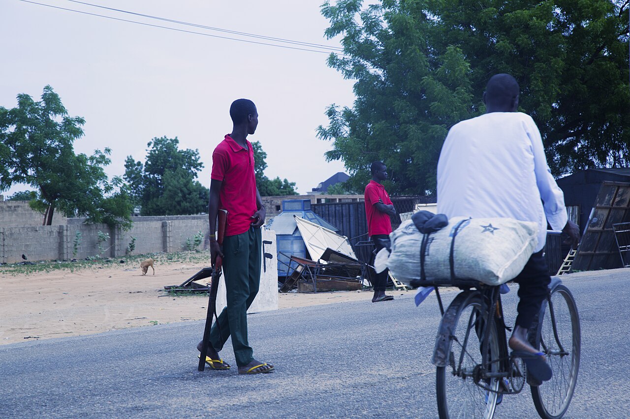 Two Civilian Joint Task Force members in red shirts stand at a roadside in Maiduguri while a man rides past on a bicycle