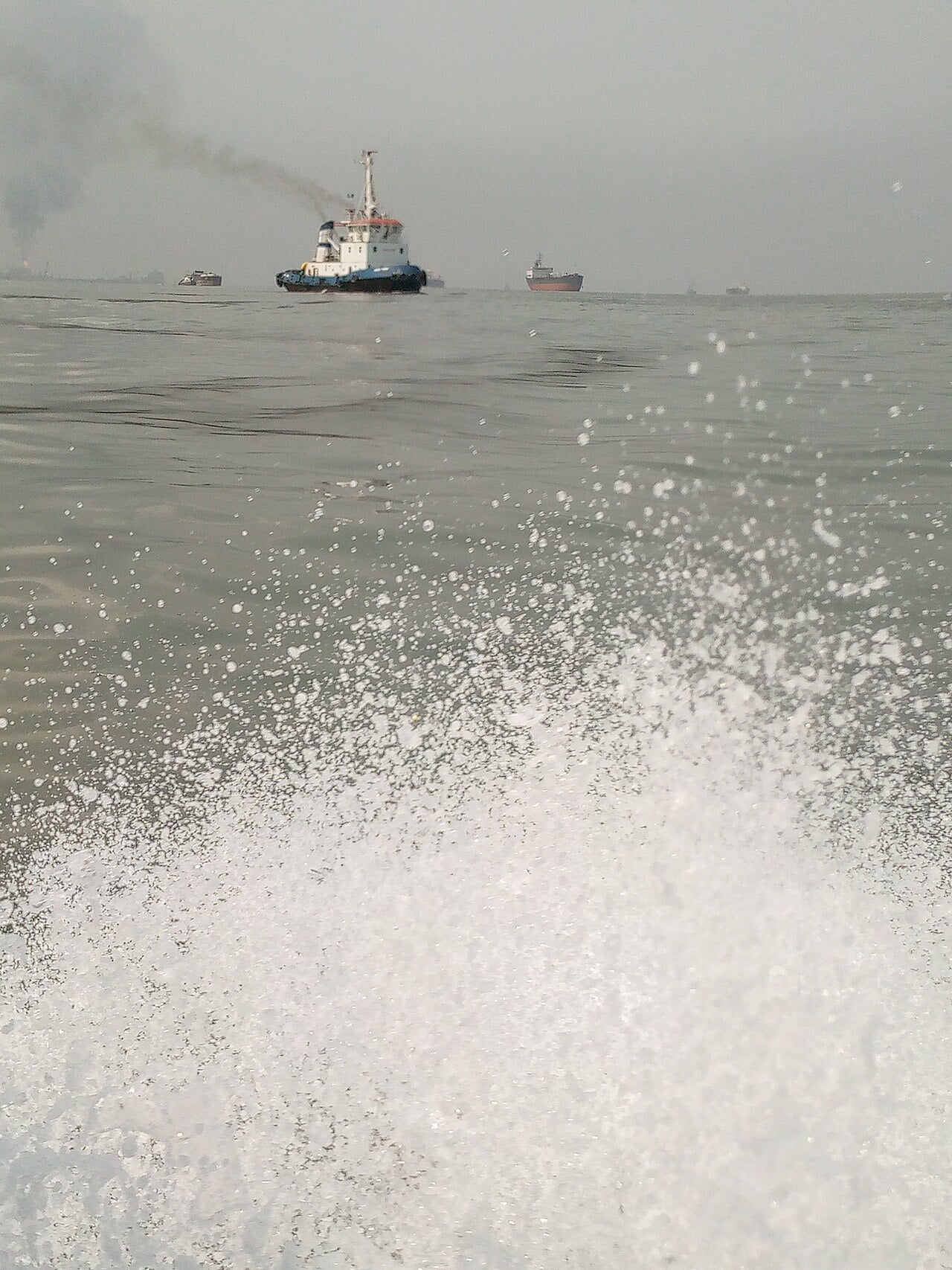 A tugboat trailing smoke and other vessels on grey water off Bonny Island, with spray from a speedboat in the foreground