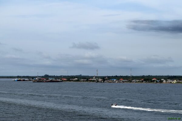 A speedboat crosses open water in front of a low stretch of Nigerian coastline with houses, moored boats and telecom masts
