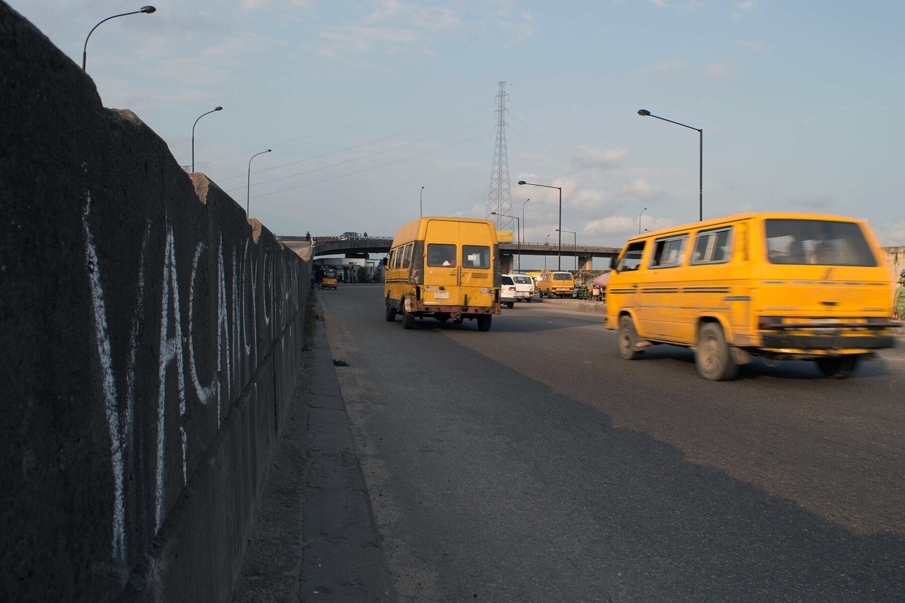 Yellow danfo minibuses driving along a road beside a graffitied wall in Lagos.