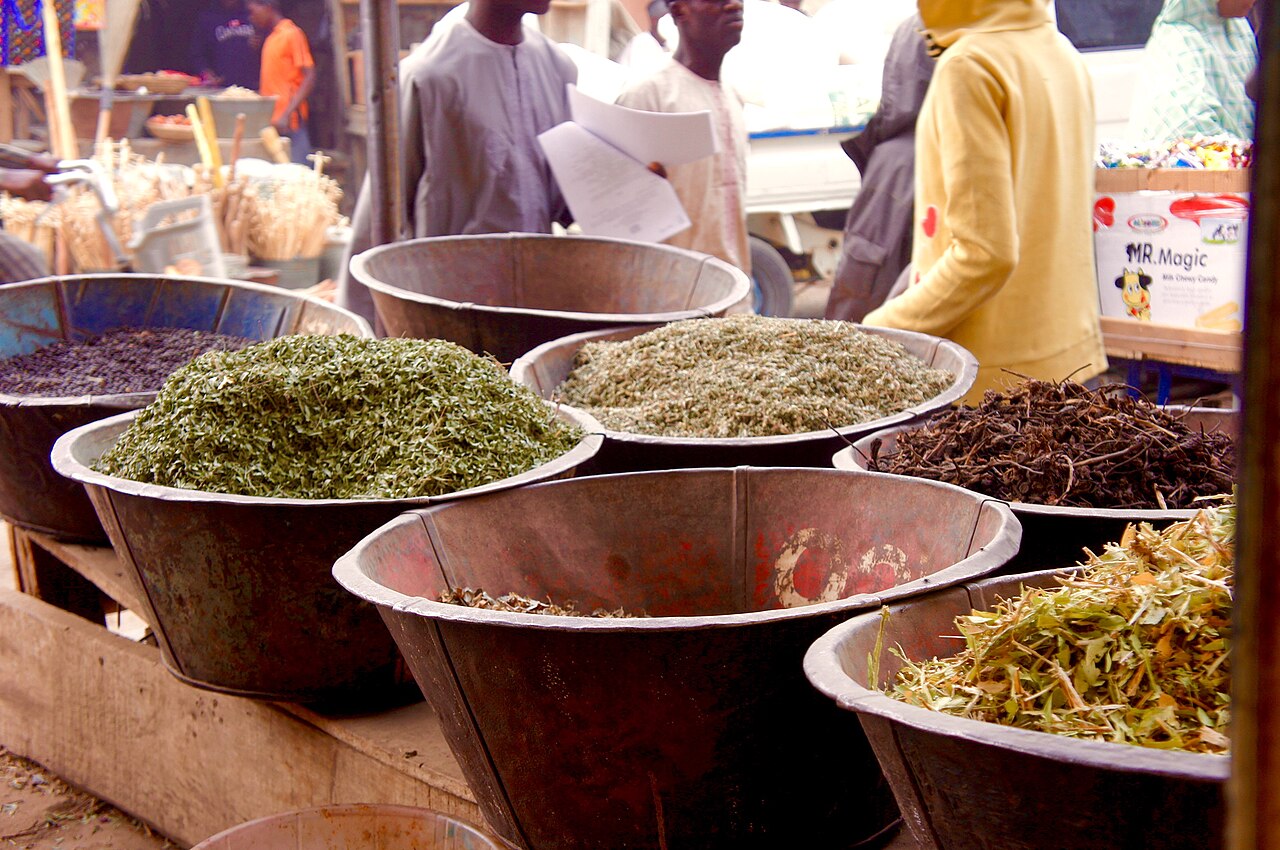 Metal basins filled with dried herbs, leaves and roots on a market stall, with shoppers behind