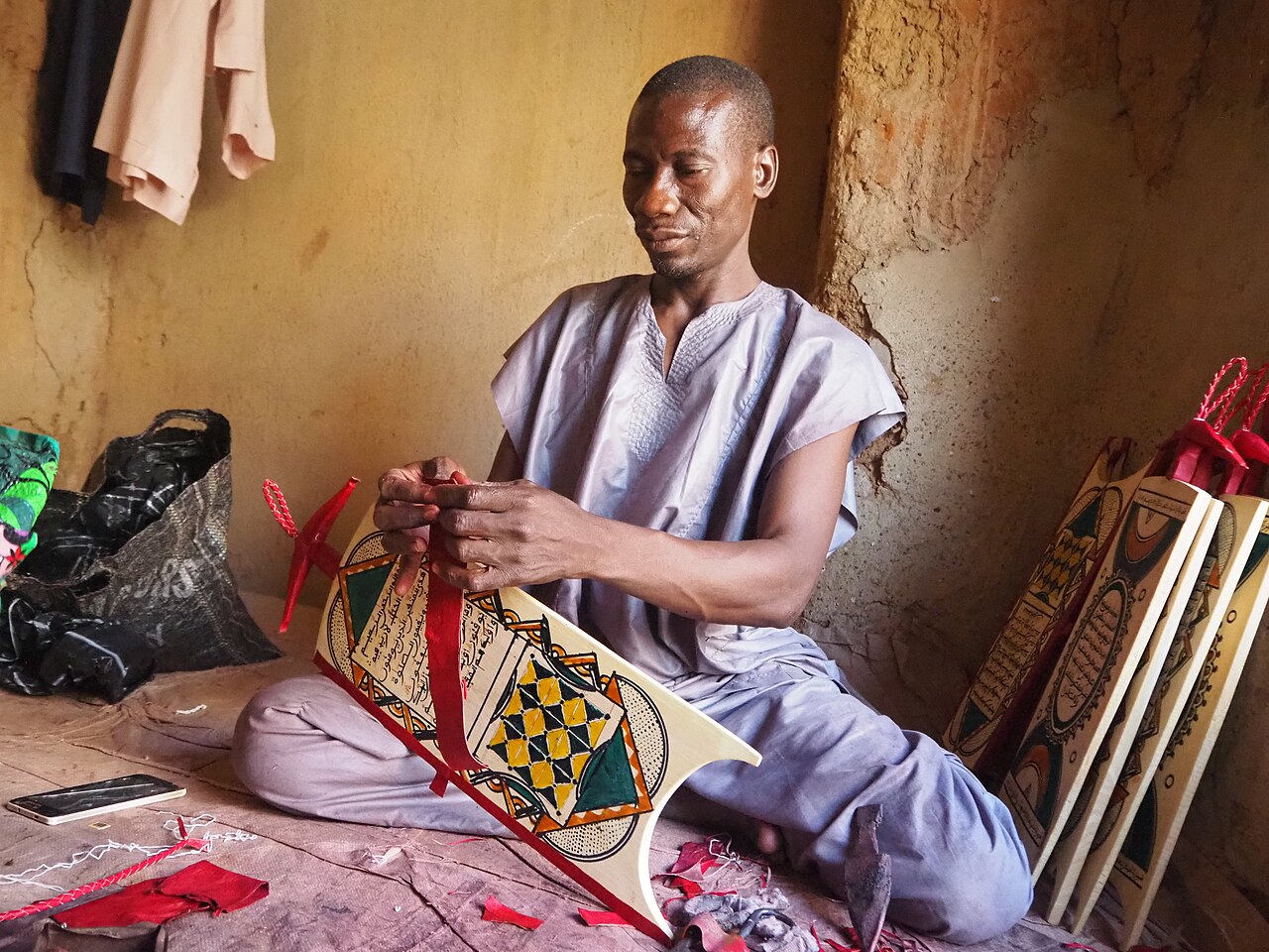A man seated on the floor ties a red strap to a painted wooden writing board with Arabic script, with more boards beside him