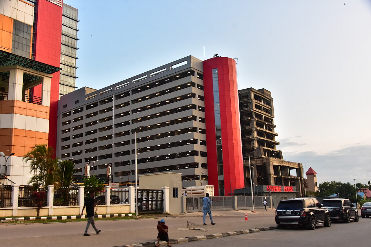 Multi-storey building with a tall red cylindrical tower beside a Revenue House sign on a street in Abuja, with parked cars and pedestrians in front