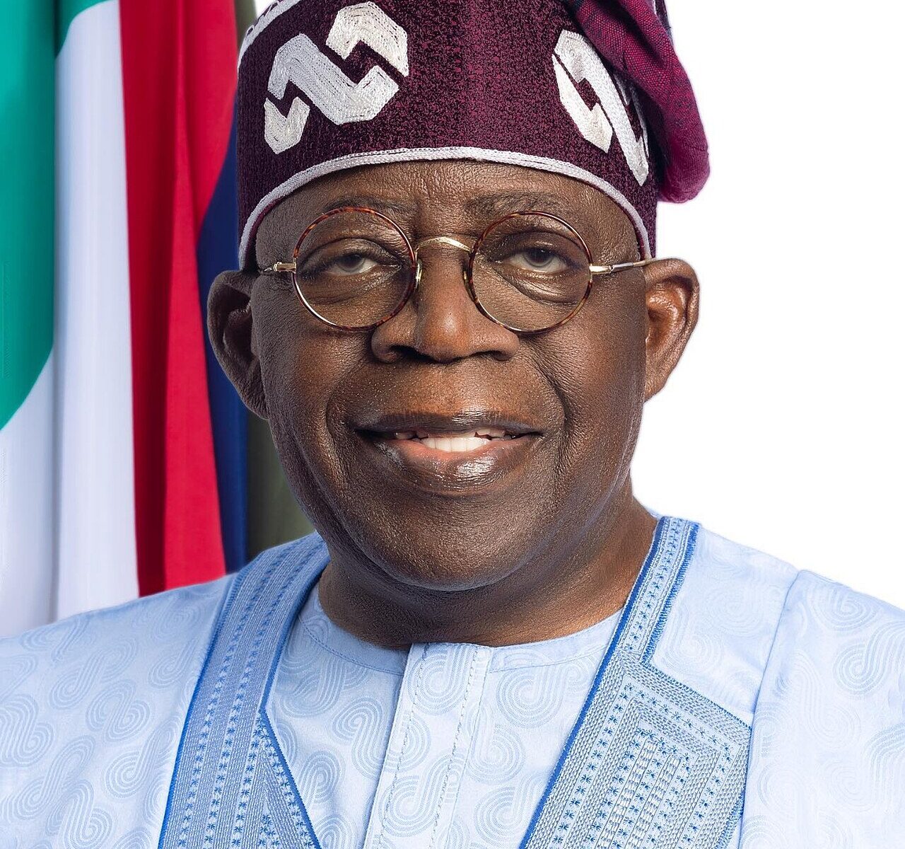 Official portrait of Nigerian President Bola Tinubu in a blue agbada and patterned cap in front of the Nigerian flag