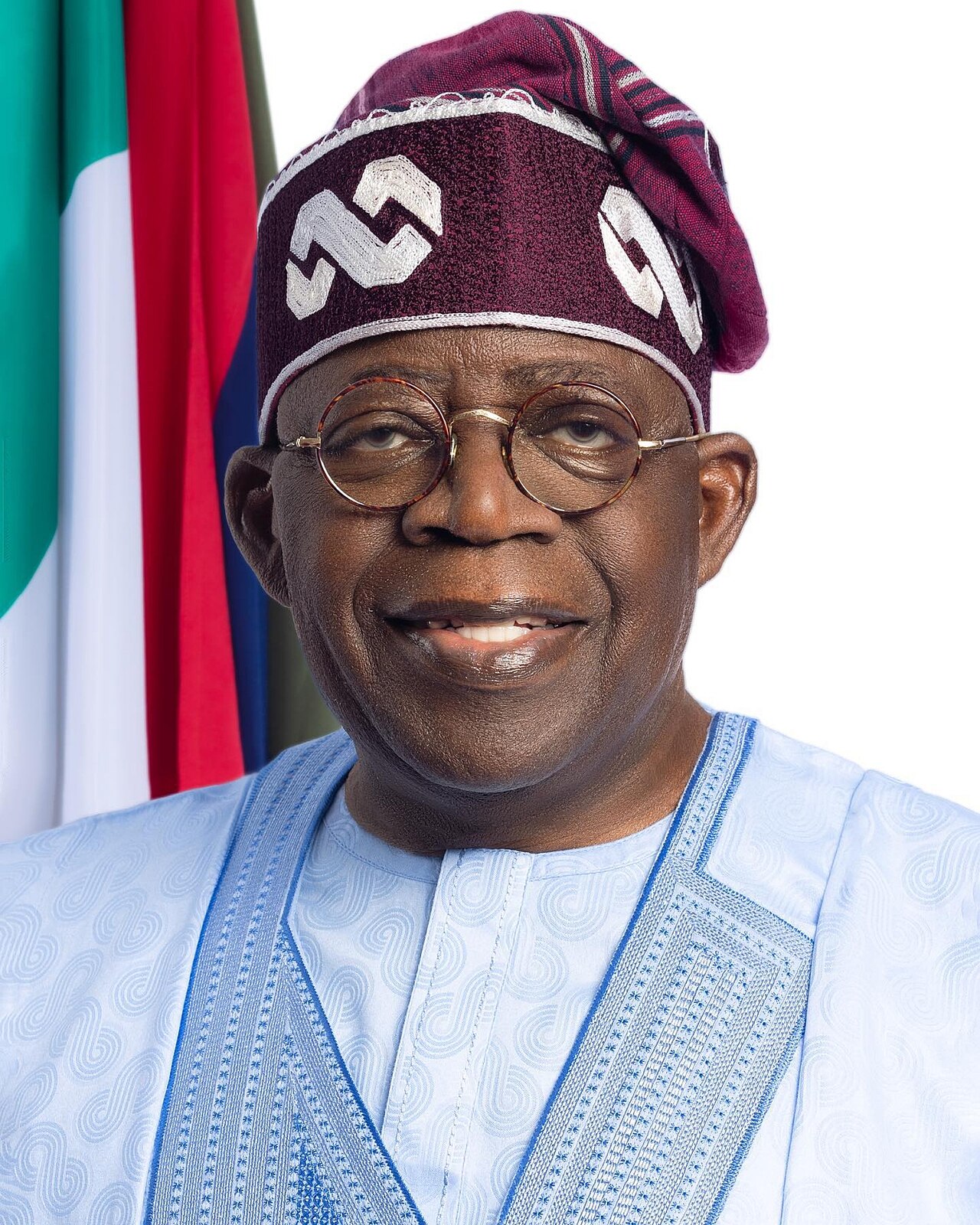 Official portrait of Nigerian President Bola Tinubu in a blue agbada and patterned cap in front of the Nigerian flag