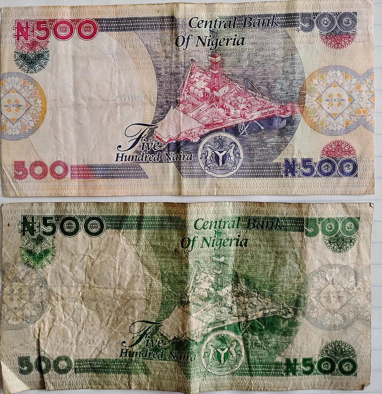 Two worn Nigerian 500 naira banknotes, one pink and blue, one green, showing the reverse side with an oil platform and the words Central Bank of Nigeria