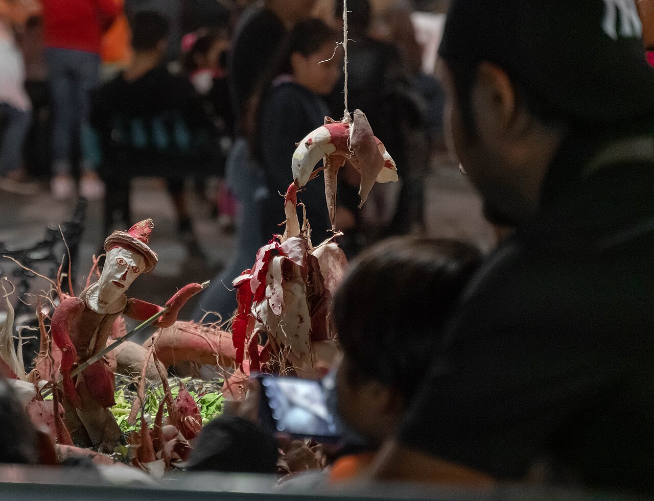 A carved radish figure in a red hat holds a stick beside a hanging radish sculpture, while visitors photograph the display at night in Oaxaca.