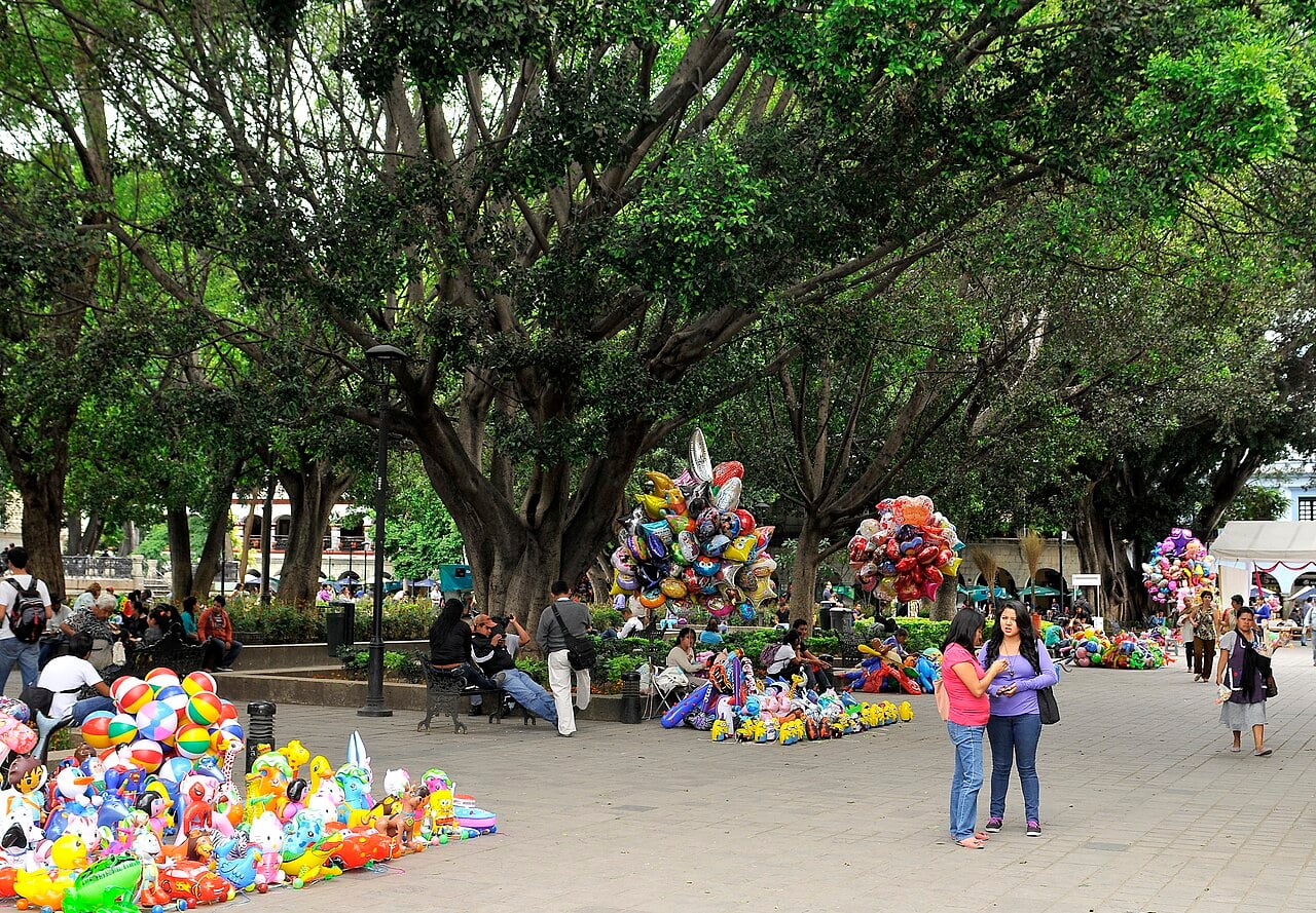 Shade trees and balloon sellers on the paved plaza of the Zócalo, the main square of Oaxaca de Juárez, with arcaded buildings behind.