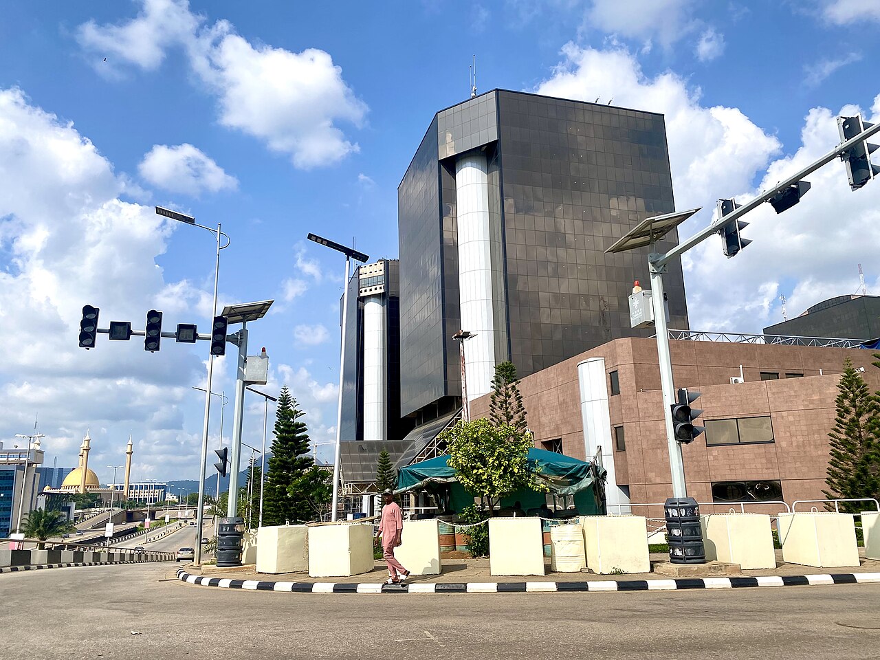 Dark glass office tower with white columns beside a road junction in Abuja, Nigeria, under a blue sky with clouds